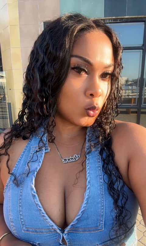 Woman with long dark curly hair wearing a blue denim halter top, posing indoors near a window with city view
